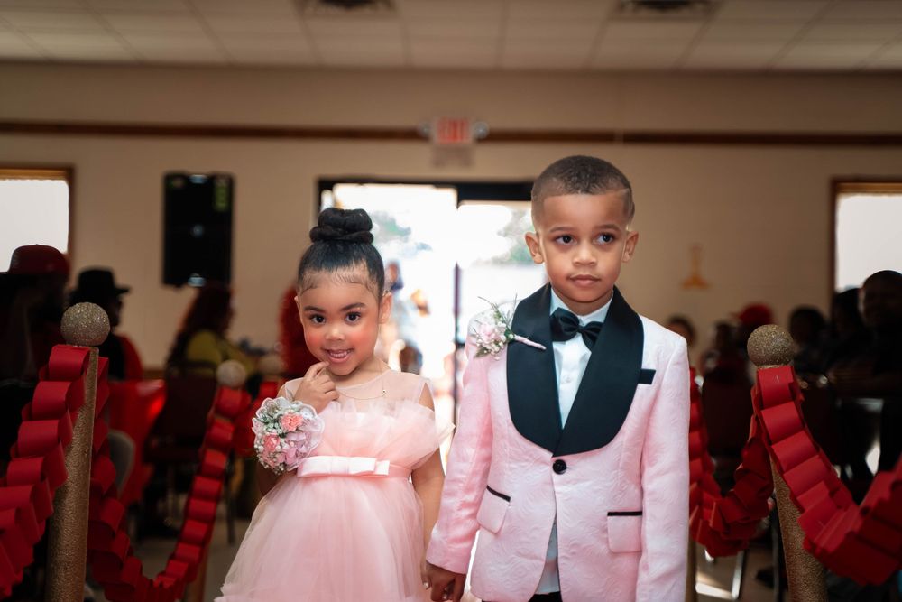 Two young children dressed formally at an event with red chairs in background.