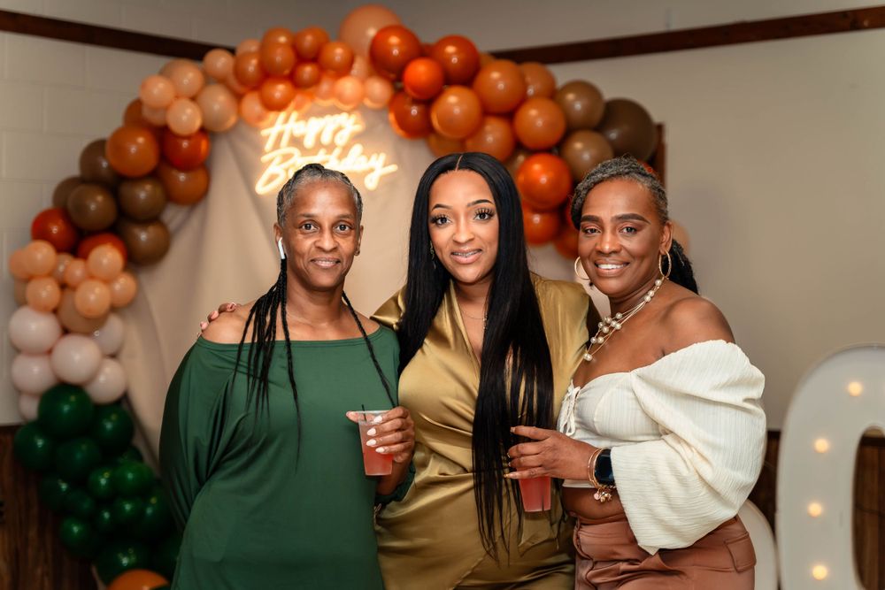 Three friends celebrating at a party under an orange and brown balloon arch decoration.
