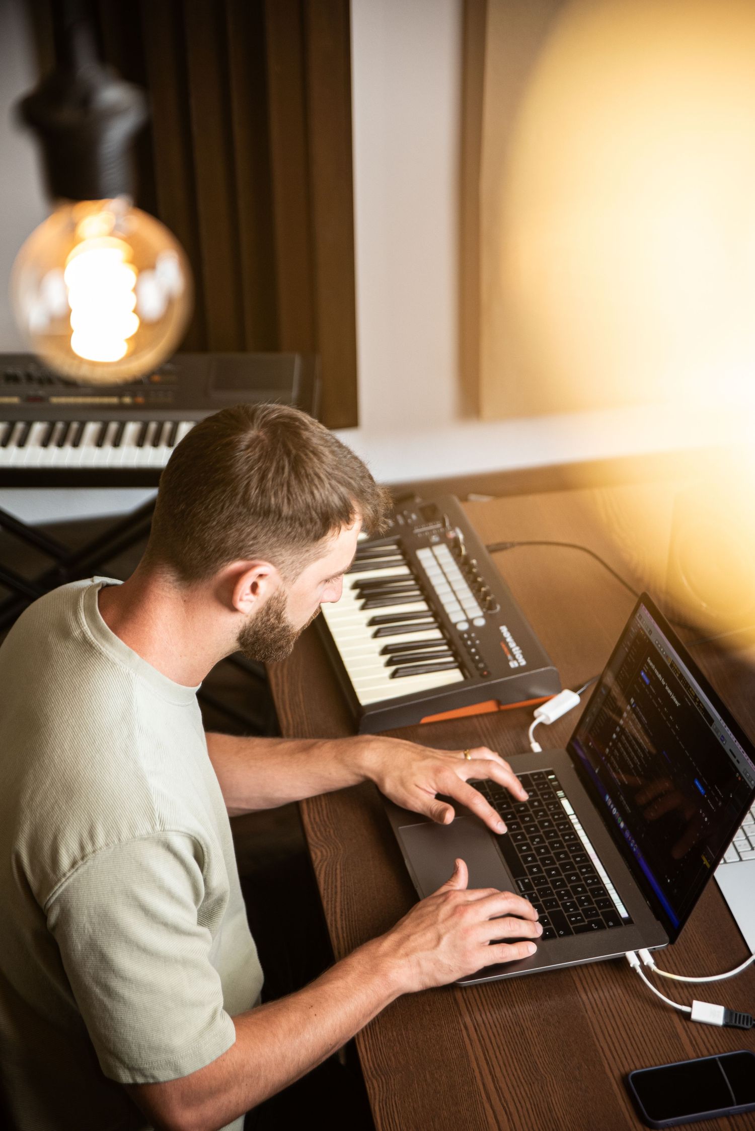 Music producer working on laptop with MIDI keyboard controllers in home studio setting.