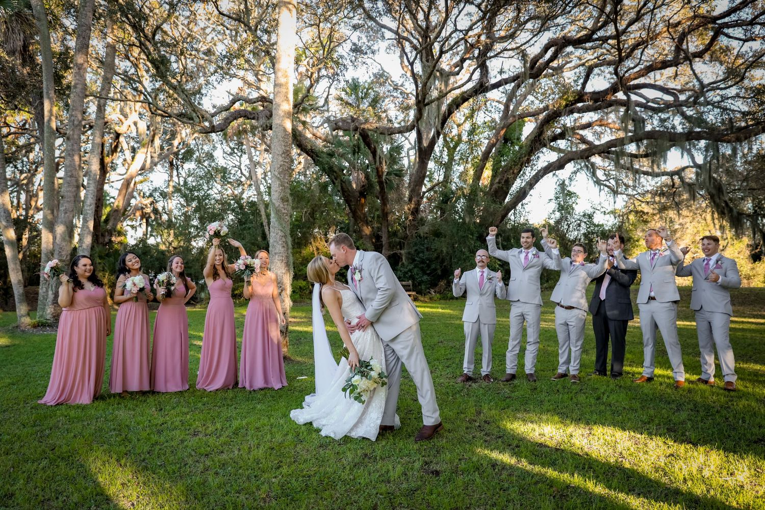 Wedding party in pink and white attire poses on lawn during golden hour.