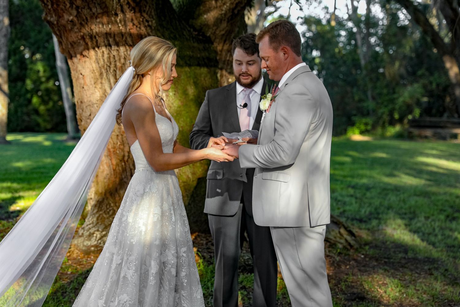 Wedding ceremony taking place outdoors under a large tree with the couple exchanging rings.