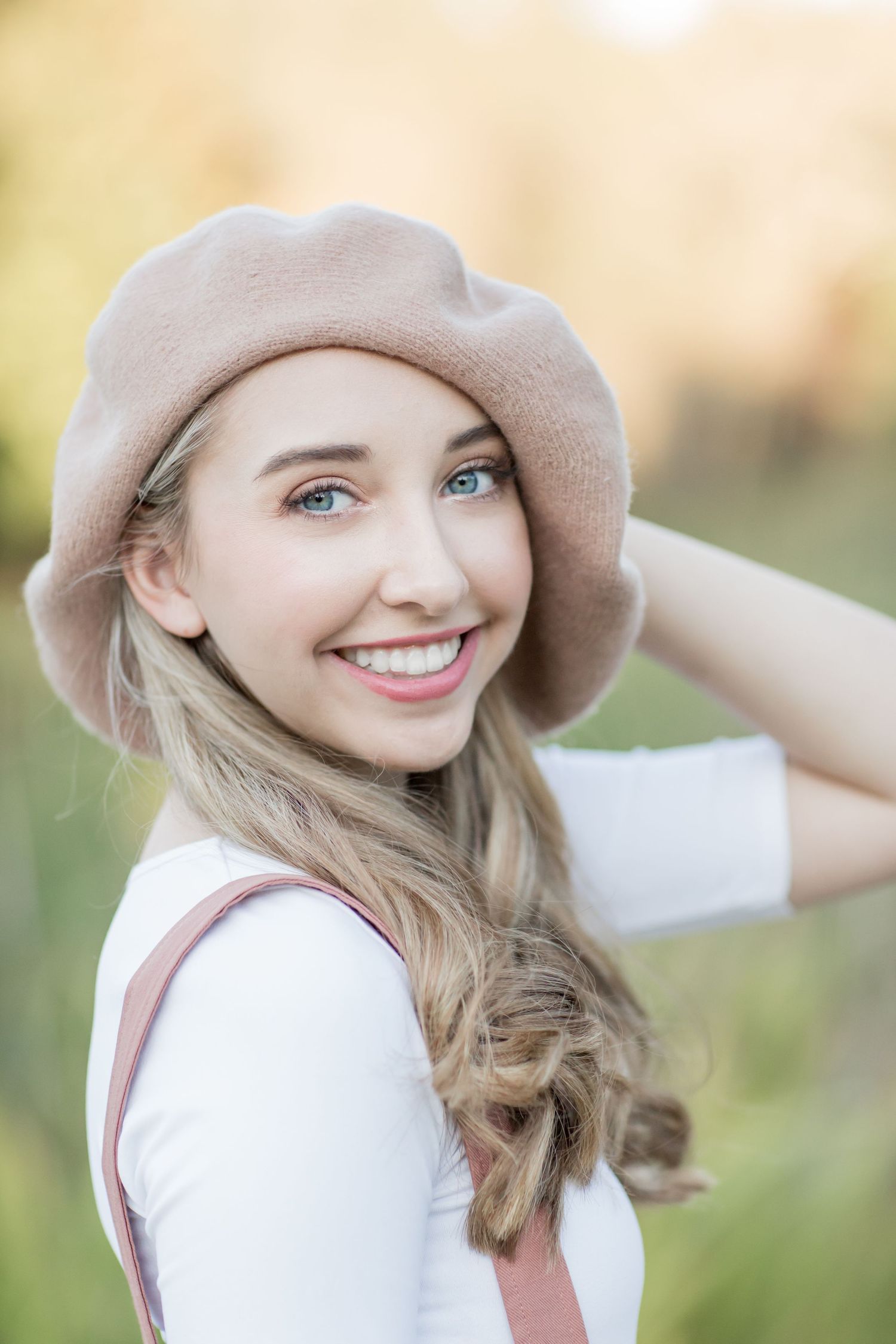 Smiling blonde young woman wearing a beige beret and white top in a sunny outdoor field.