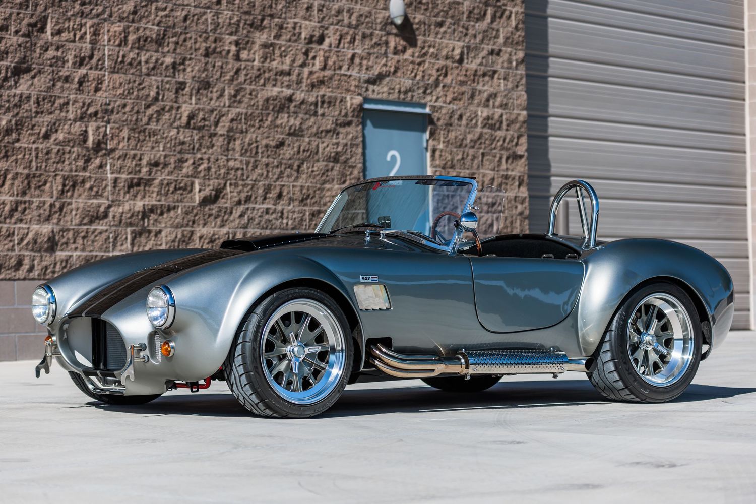 Classic silver Shelby Cobra roadster with racing stripes parked against textured wall.