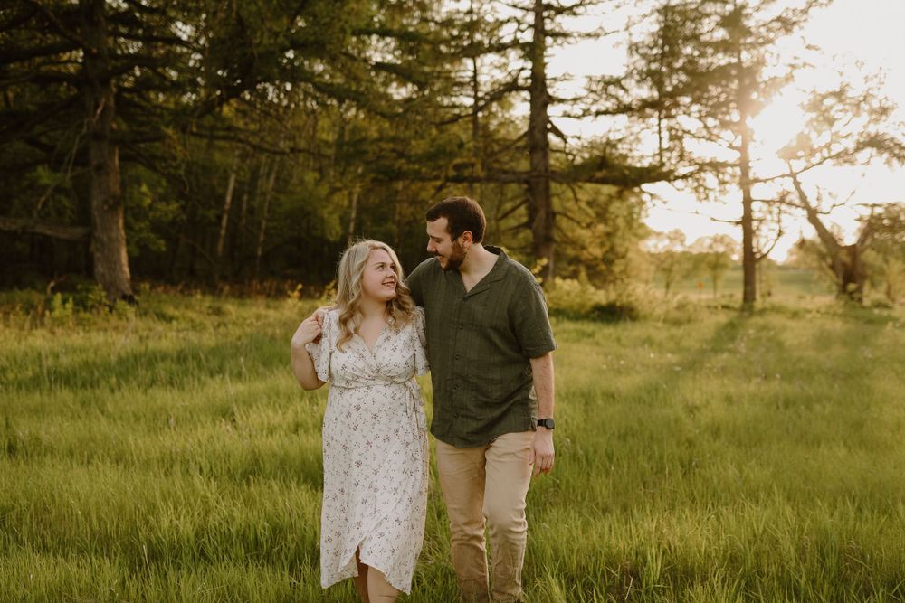 Couple walking together through sunlit meadow with pine trees in background.