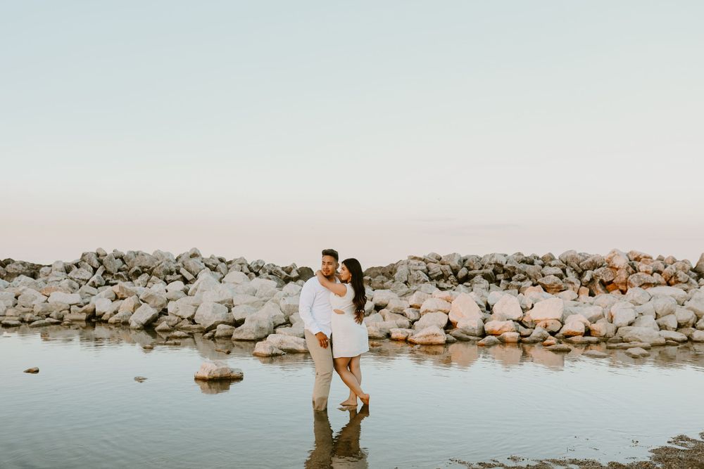 Couple embracing while standing in shallow water near rocky shoreline at sunset.