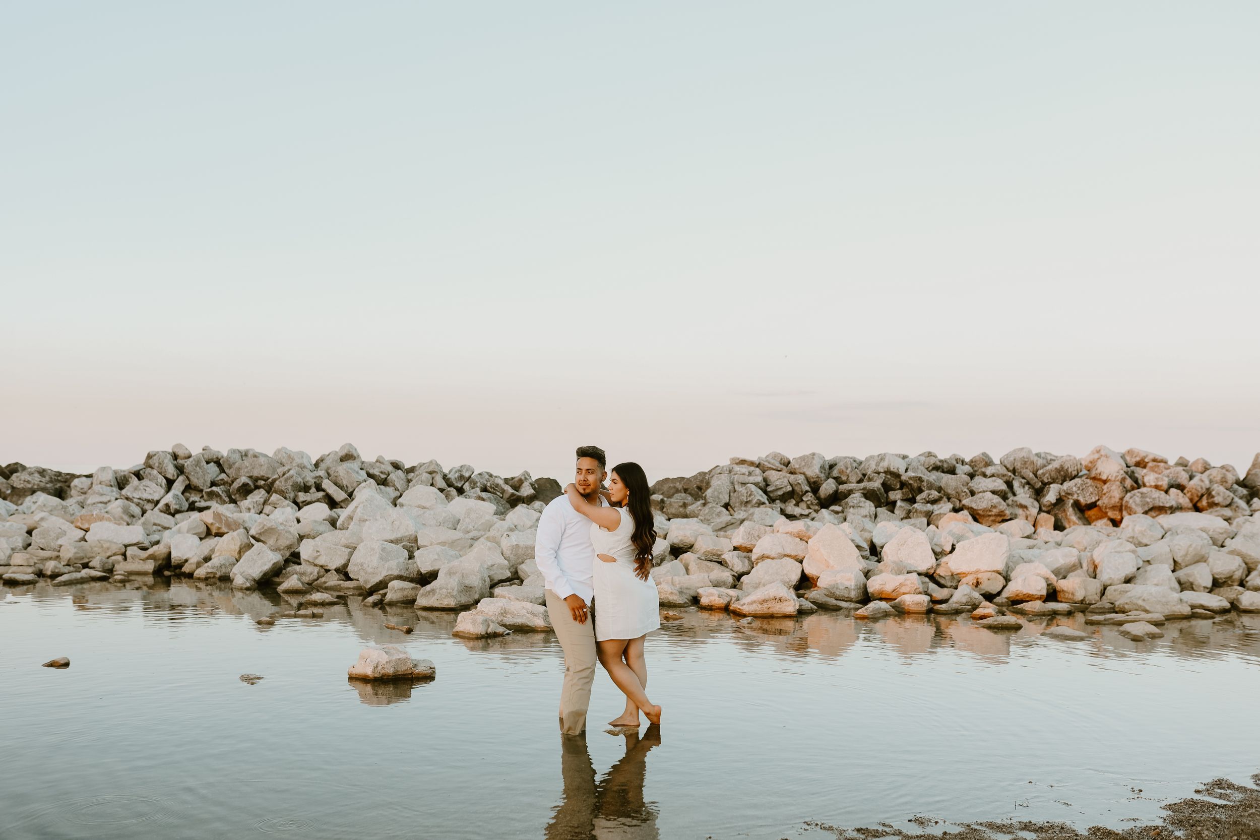 Couple embracing while standing in shallow water near rocky shoreline at sunset.