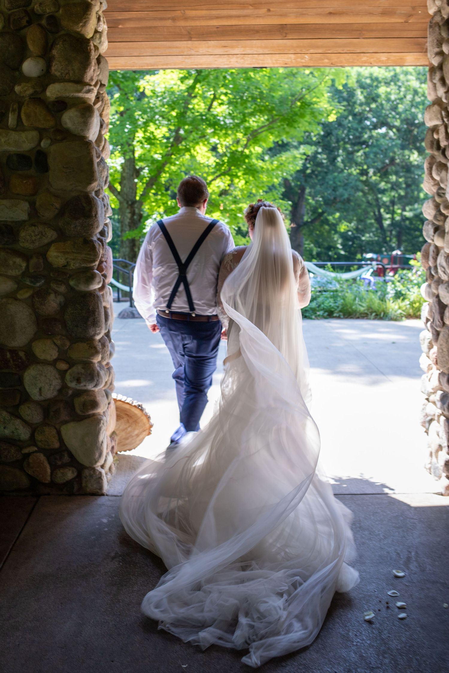 Bride and groom walking hand-in-hand through a rustic stone archway into lush greenery.