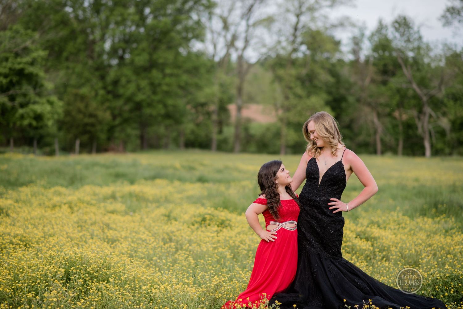 Two figures in formal dresses, one black and one red, pose together in a field of yellow flowers.