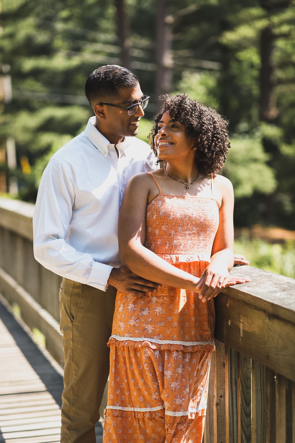 Couple in orange dress and khakis share tender moments on a wooden bridge in nature.