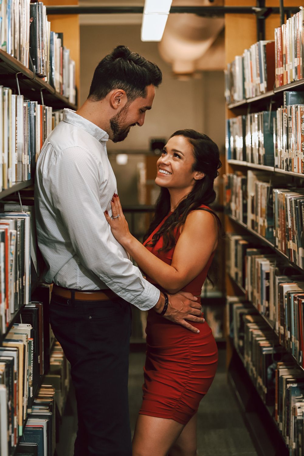 Romantic couple embracing between library bookshelves in casual formal attire.