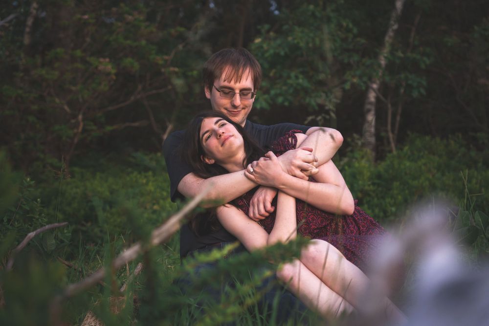 A couple embraces in a wooded area with natural lighting filtering through trees.