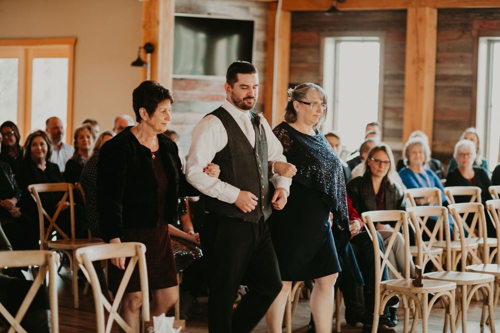 Groom is escorted down the aisle by two women in a warmly lit rustic barn wedding ceremony.
