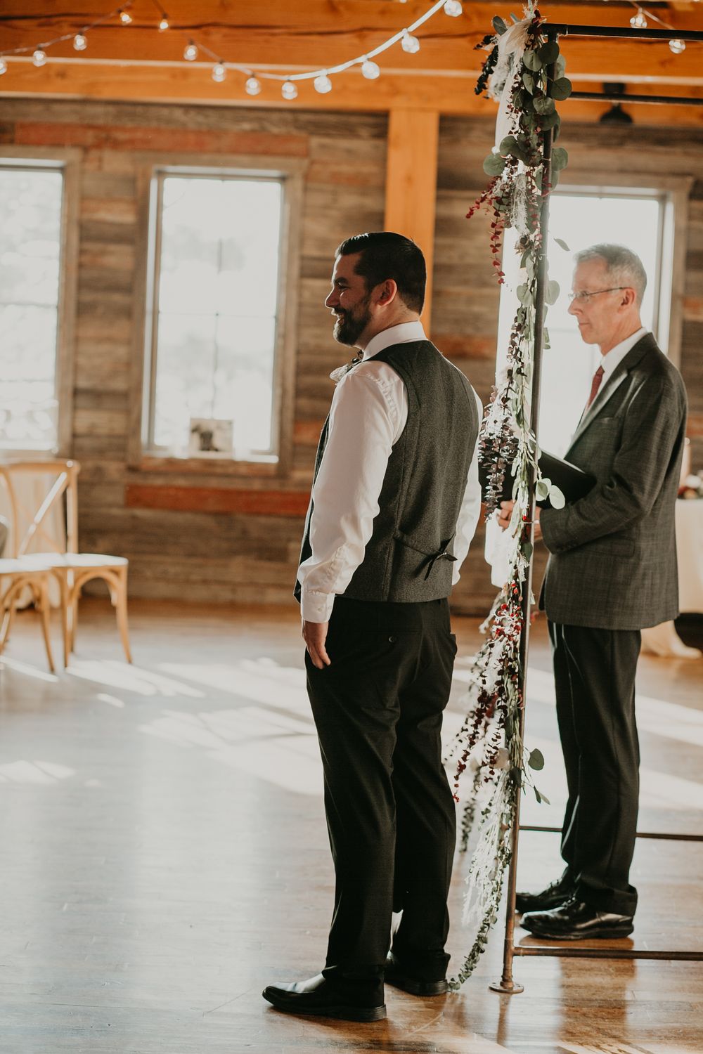 Groom in grey vest stands at floral-adorned altar with officiant inside a rustic wooden barn venue.