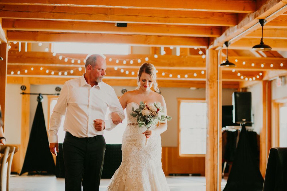 Bride in lace gown walks down the aisle with her father in a charming rustic barn wedding venue.
