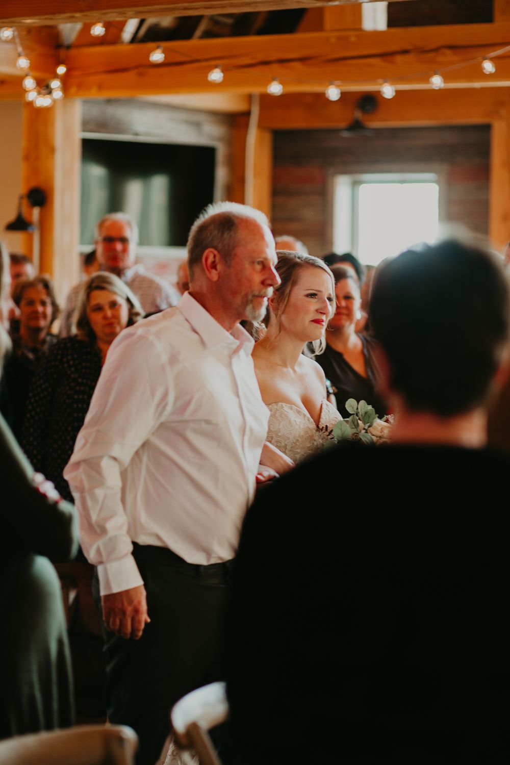 Bride in lace gown walks down the aisle with her father toward the groom in a rustic barn venue.