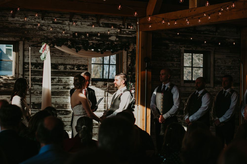 Bride and groom hold hands during their candlelit rustic barn wedding ceremony with wedding party.