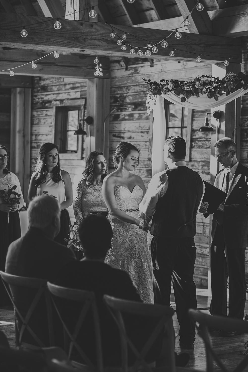 Black and white photo of bride reading vows at rustic barn wedding ceremony with bridesmaids.