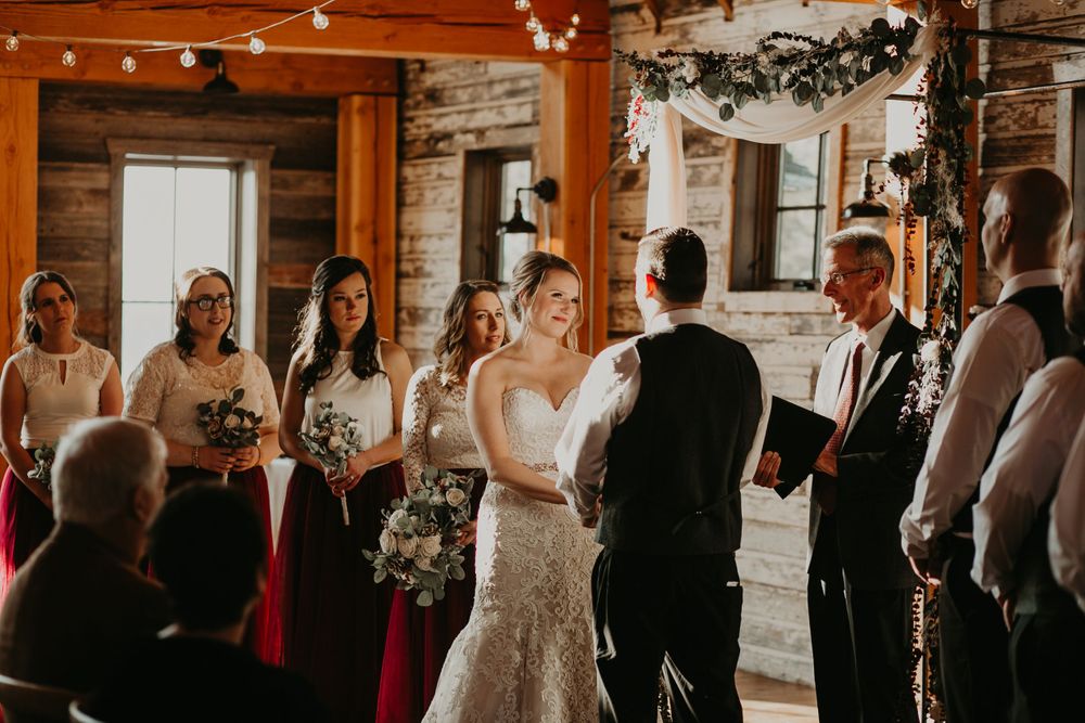 Smiling bride exchanging vows with groom at rustic barn wedding, bridesmaids in red dresses.