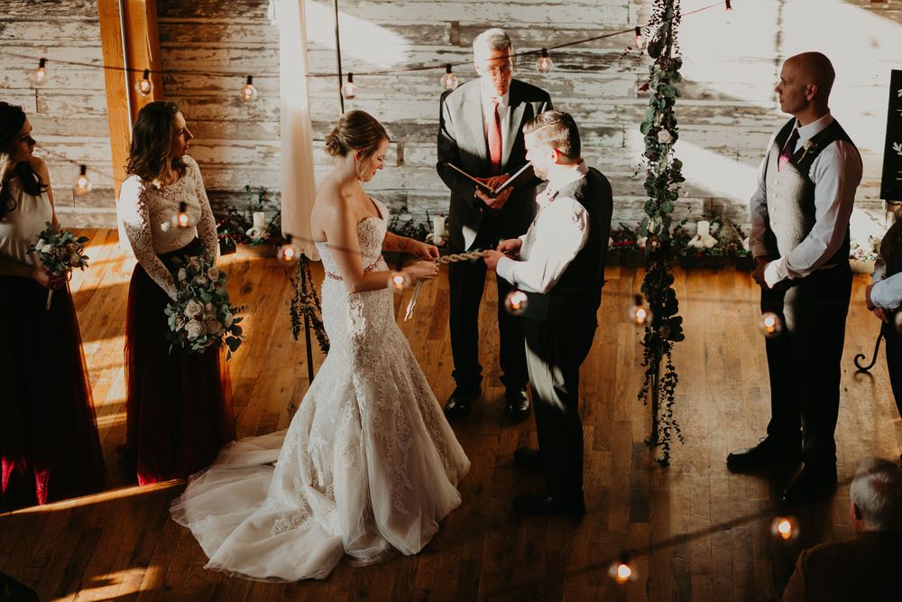 Bride and groom holding hands during rustic barn wedding ceremony with officiant and wedding party.