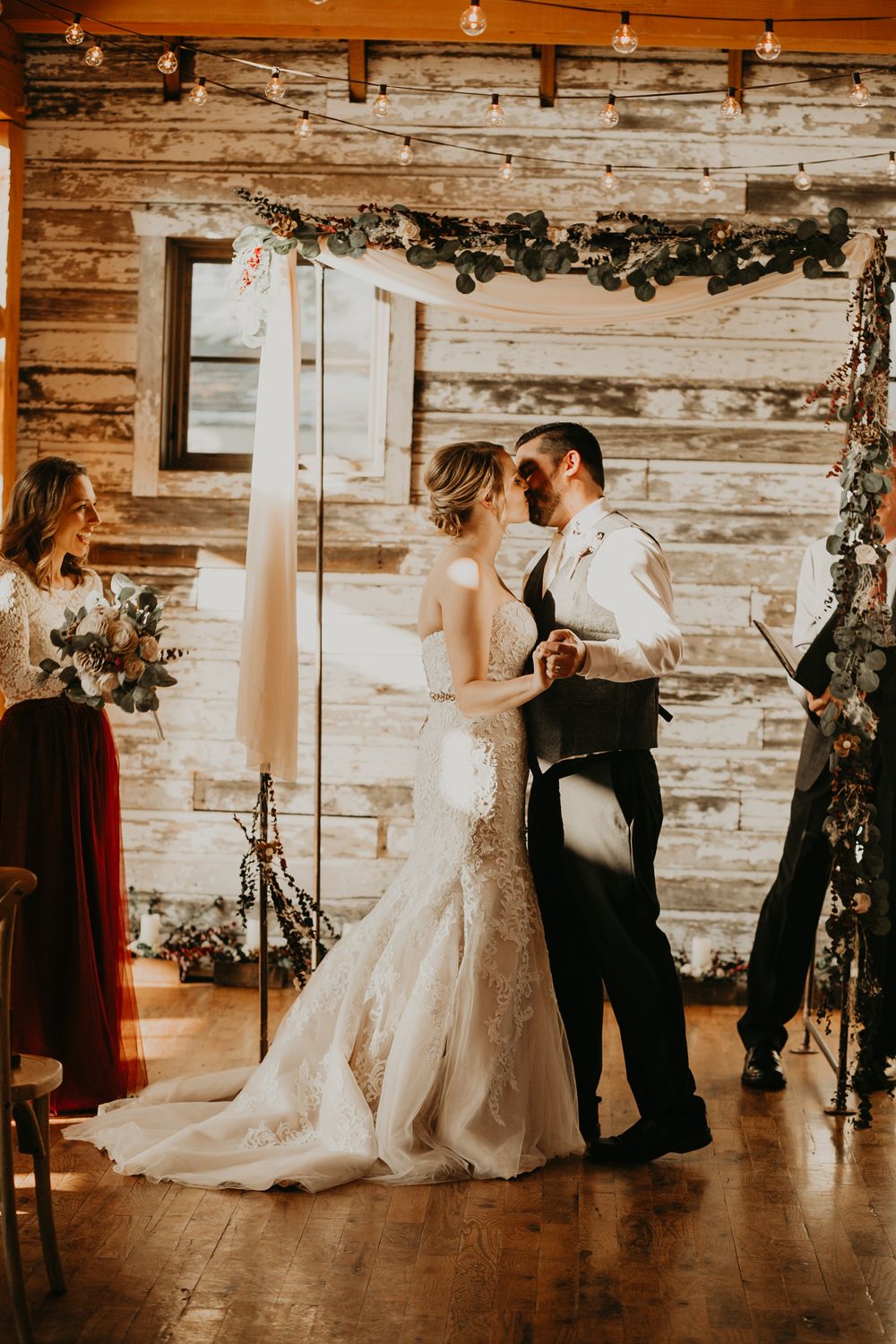 Bride and groom sharing first kiss at rustic barn wedding altar adorned with greenery garland.