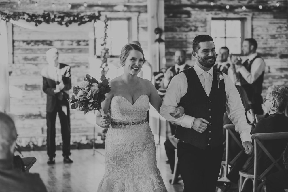 Black and white photo of joyful newlyweds walking down the aisle after barn wedding ceremony.
