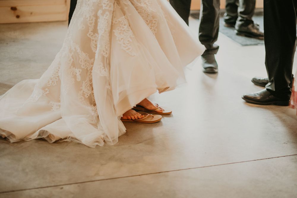 Bride in lace wedding gown wearing gold sandals on a concrete floor at rustic wedding.