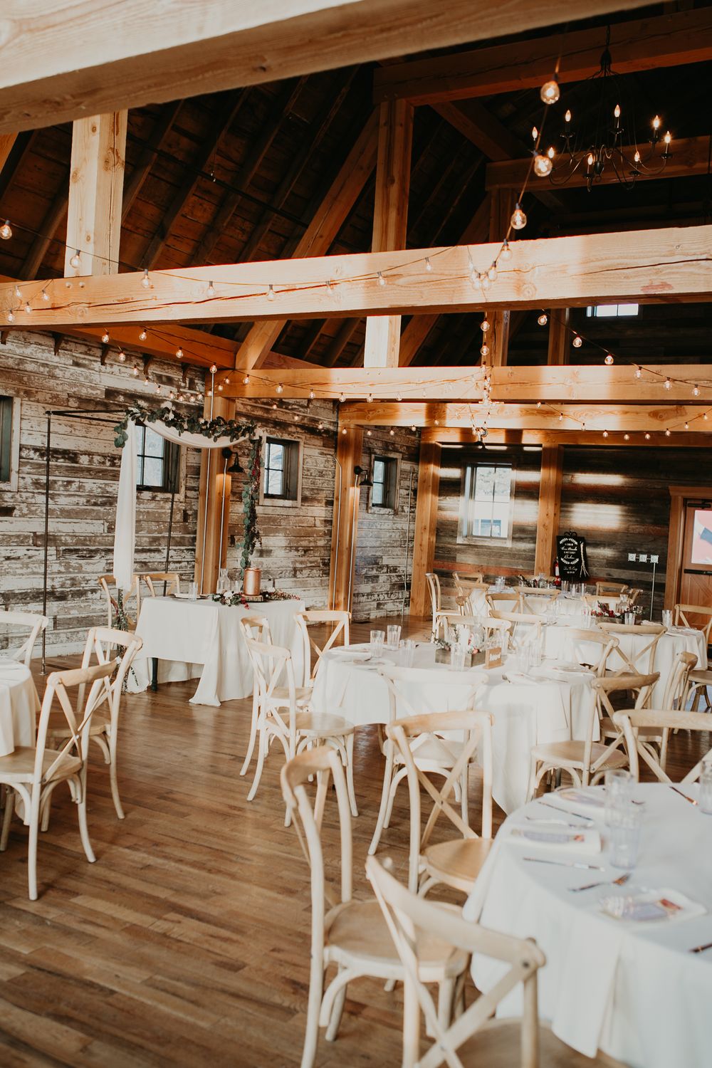 Rustic barn wedding reception venue with wooden beams, string lights, and white round tables.