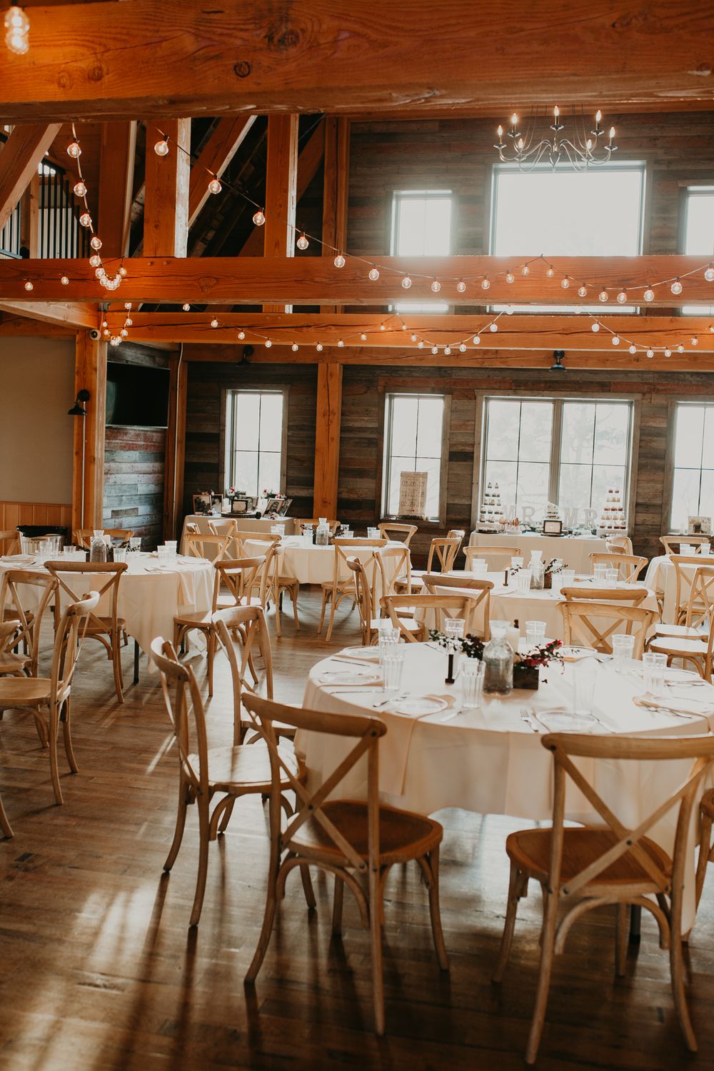 Warm rustic barn wedding reception hall with wooden chairs, string lights, and round tables.