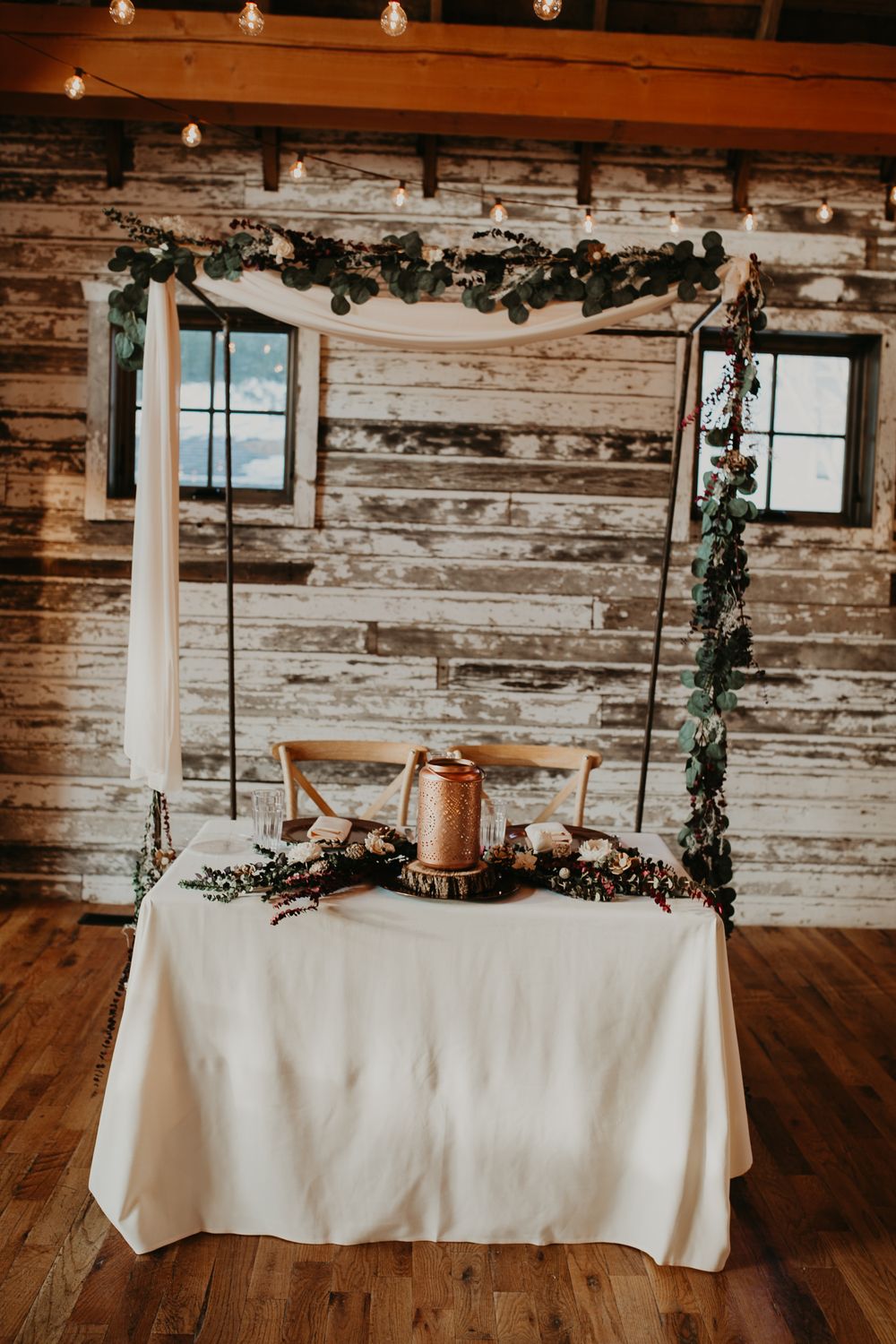 Rustic sweetheart table with greenery arch, white draping, and copper lantern centerpiece.