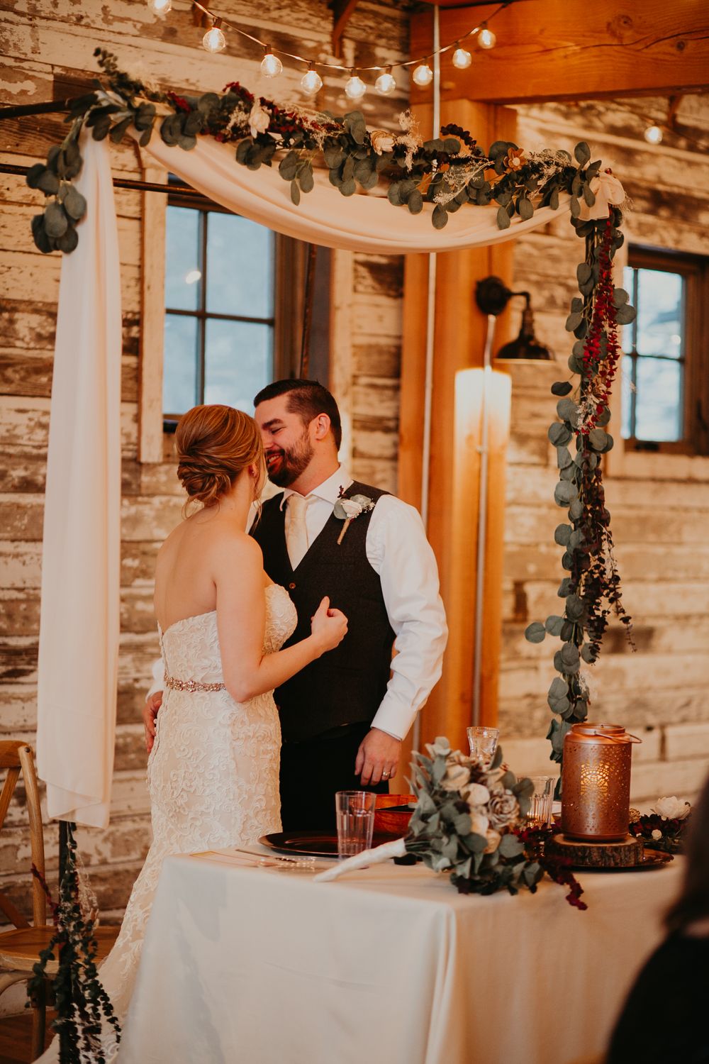 Bride and groom share a romantic moment at rustic barn wedding reception under eucalyptus garland arch with string lights.