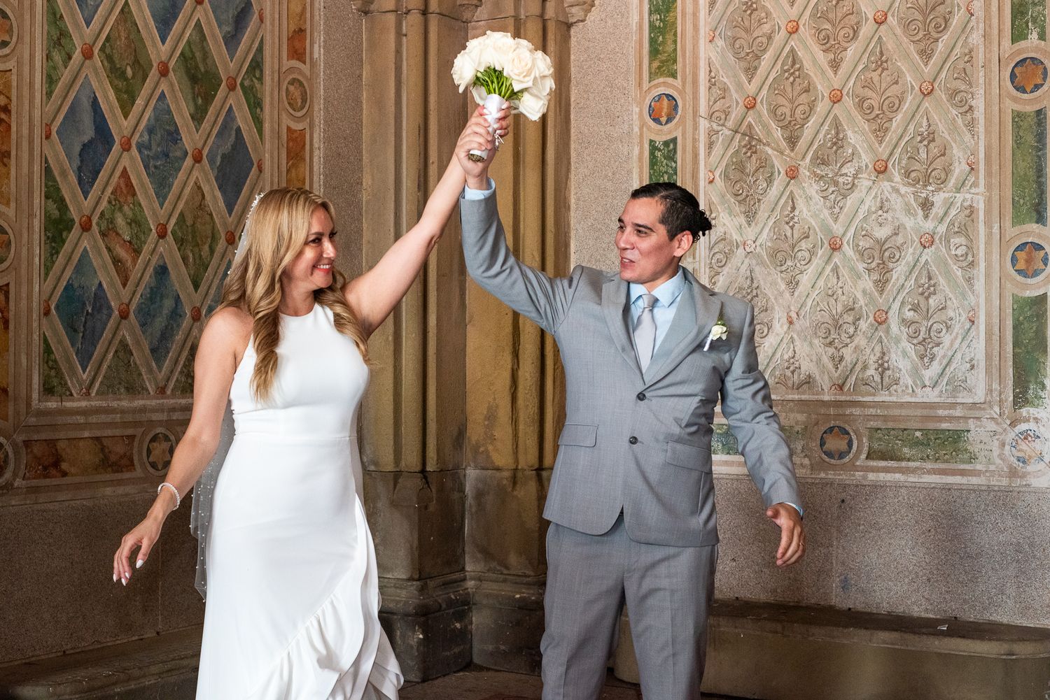 Joyful wedding couple celebrates with raised bouquet against decorative architectural background.