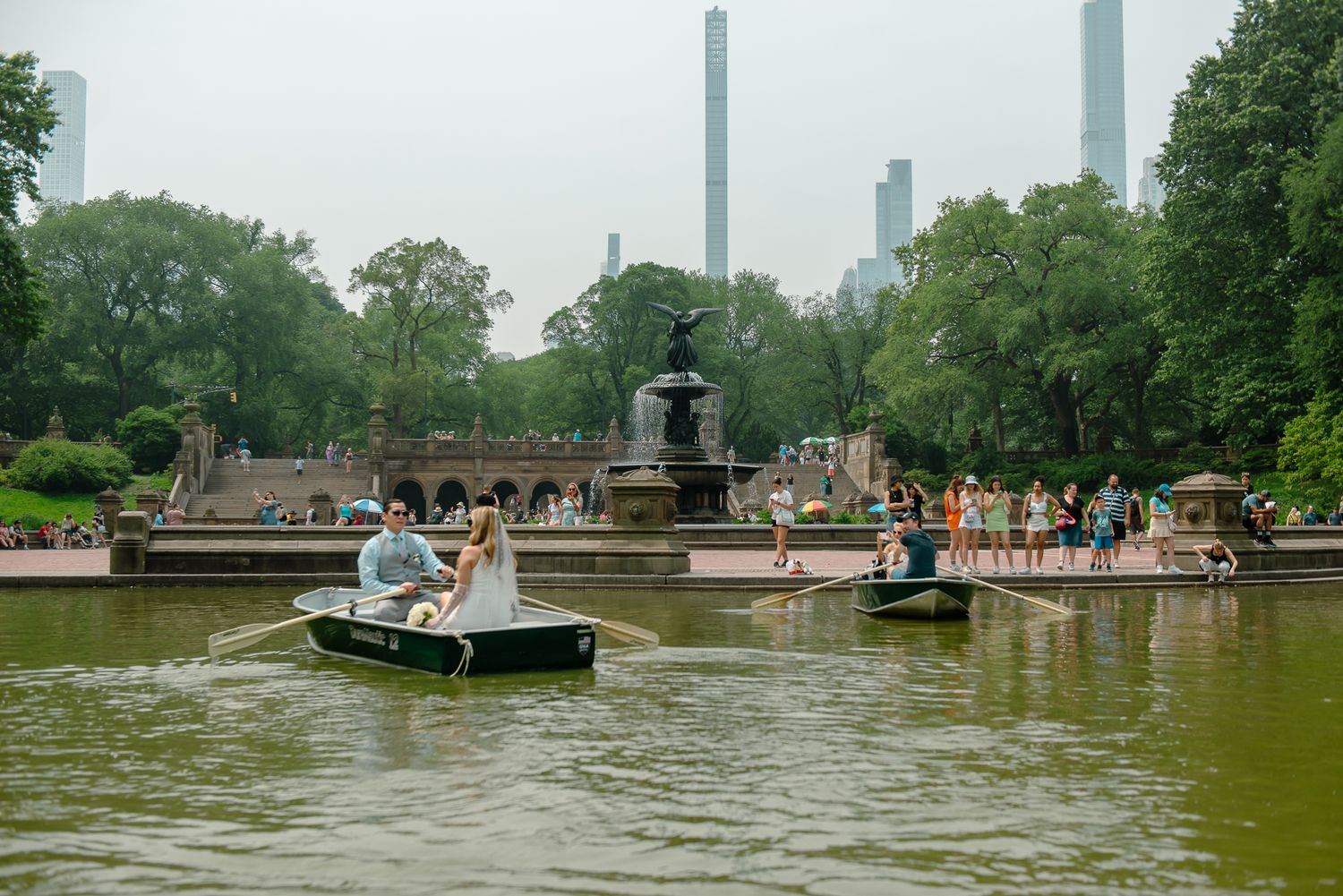 Wide angle view of rowboats on Central Park lake with city buildings visible in distance.