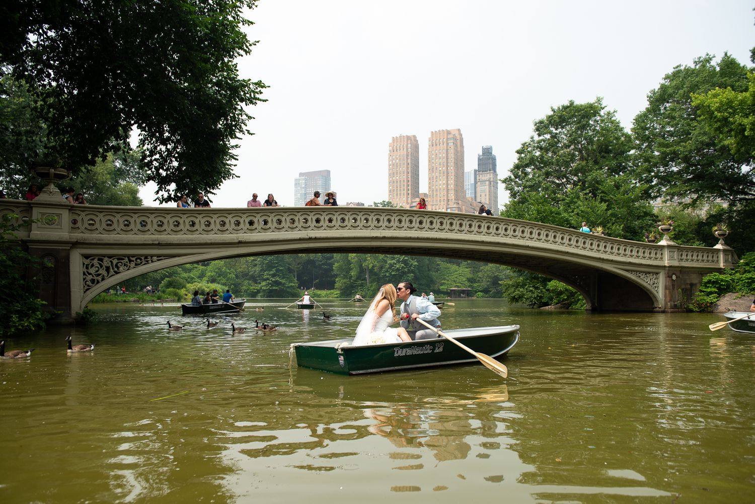 Wedding party in rowboats gliding across Central Park lake with bridge in background.