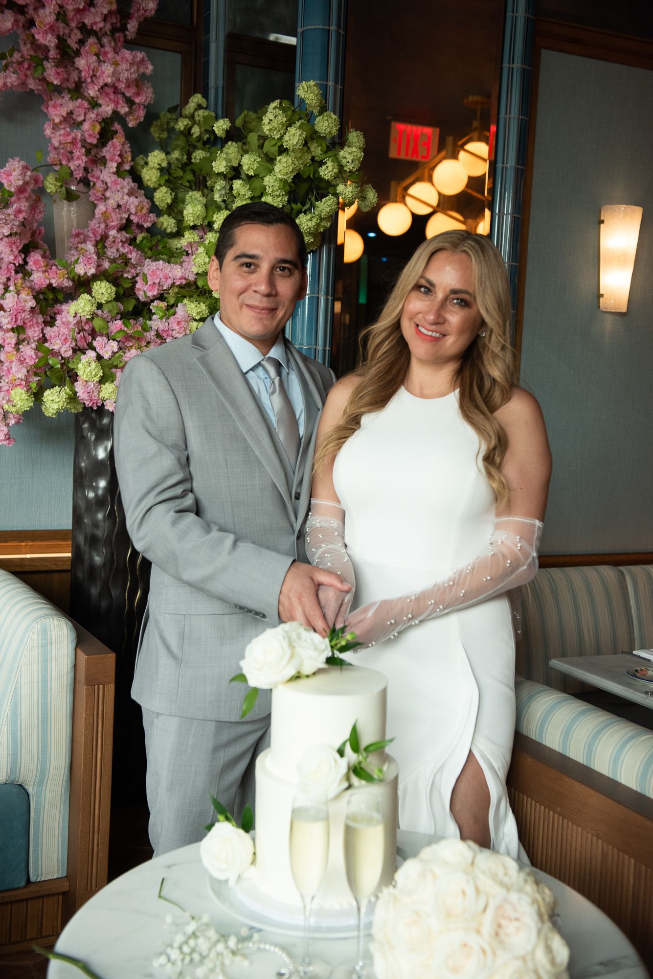 A couple in wedding attire cuts their elegant white two-tier cake decorated with roses at an intimate reception.