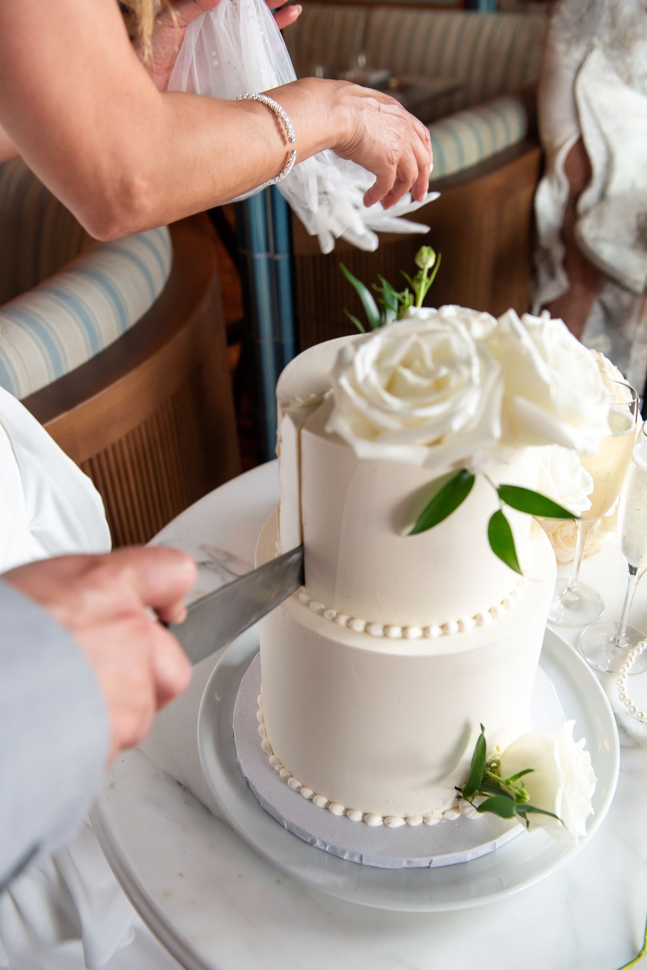Close-up of hands cutting into a white two-tier wedding cake adorned with fresh roses and greenery.