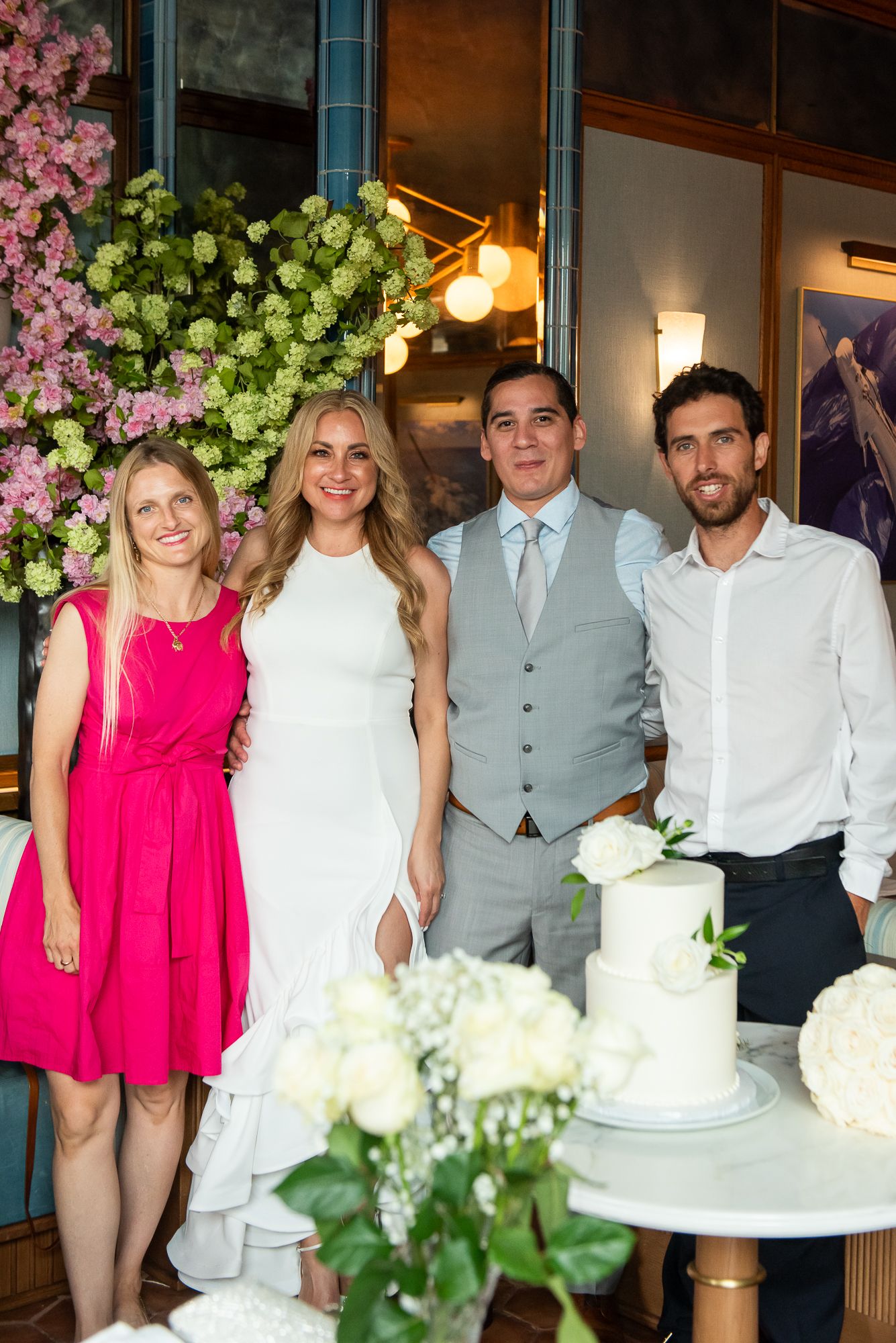 Wedding group photo with guests in pink and grey attire posing by a white cake.