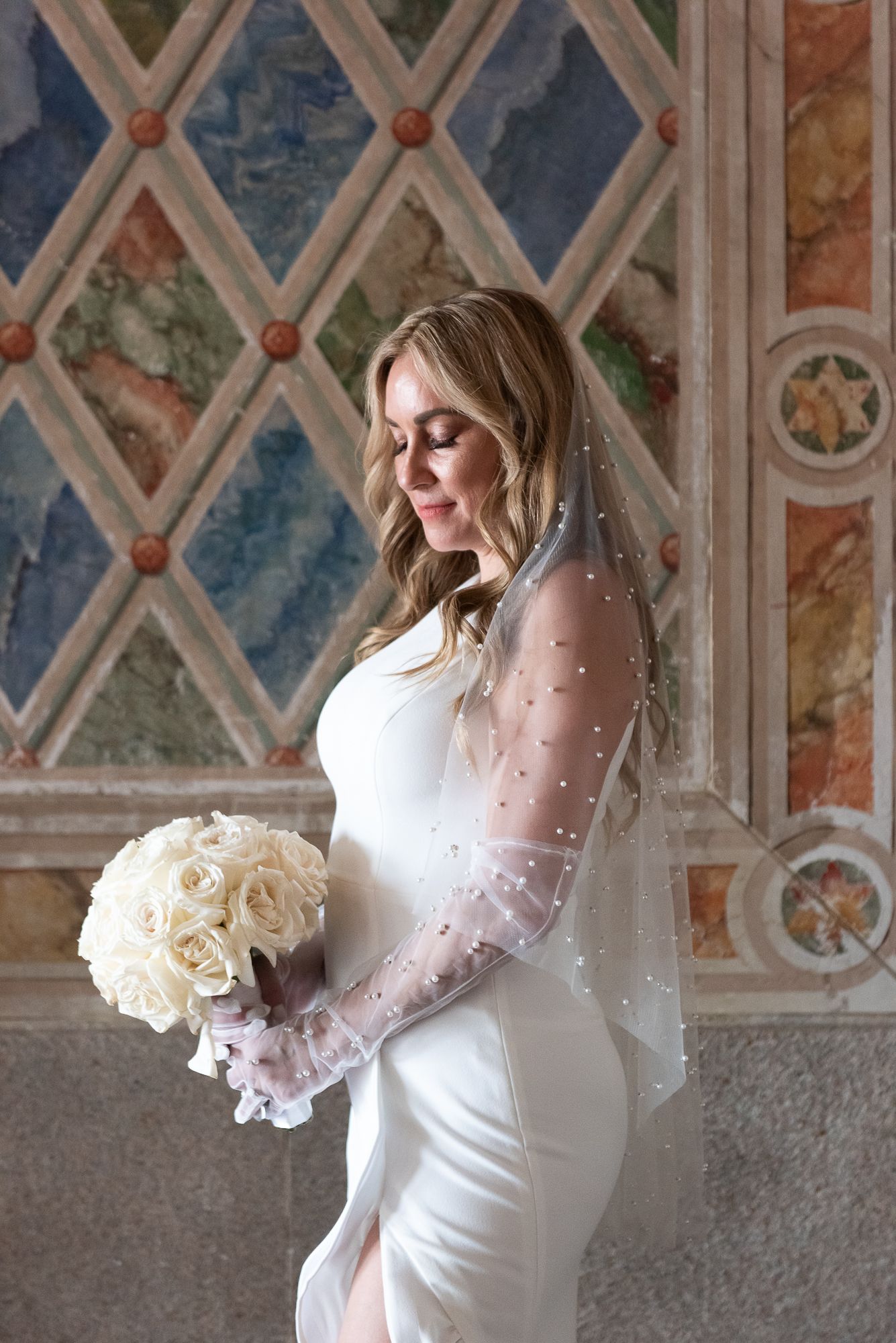 Elegant white wedding dress with sheer sleeves and white roses bouquet against decorative wall.