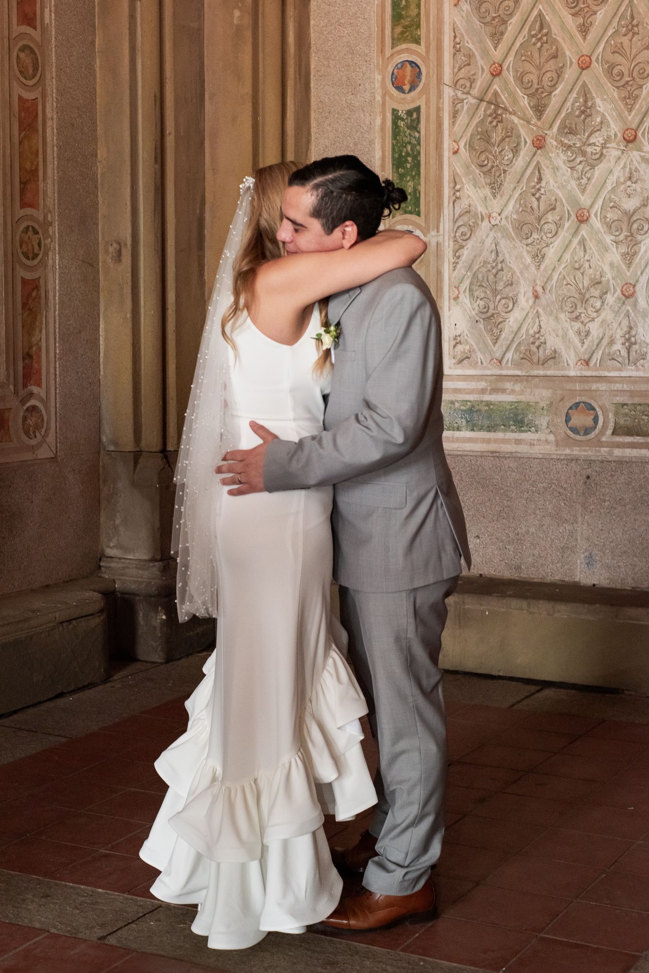 A romantic embrace between newlyweds against an ornate patterned wall in a historic venue.
