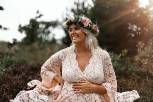 Ethereal outdoor maternity portrait with flowing white lace dress and floral crown in natural setting.