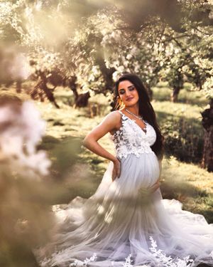 Ethereal maternity portrait in white lace gown with flowering trees and golden sunlight.