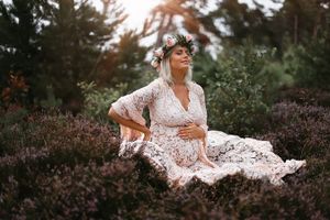 A dreamy maternity photo in a white lace dress surrounded by purple heather and evergreen trees at sunset.