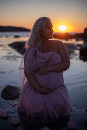 Dreamy sunset maternity photo on rocky beach with flowing pink dress and golden light reflecting on water.