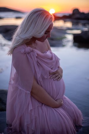 Ethereal maternity portrait at sunset with pink flowing dress against ocean backdrop.