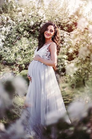 Dreamy spring maternity photo featuring flowing white lace dress amid blooming trees.