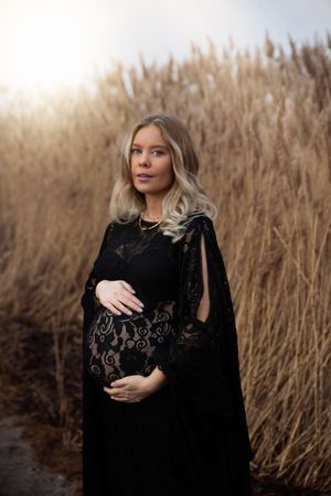 Dramatic maternity portrait in black lace dress against tall wheat field at golden hour.