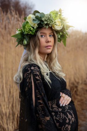Elegant maternity portrait with green floral crown and black lace dress against golden wheat field.