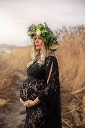 Expectant woman in black lace dress wearing floral crown poses in field of tall grass at sunset.