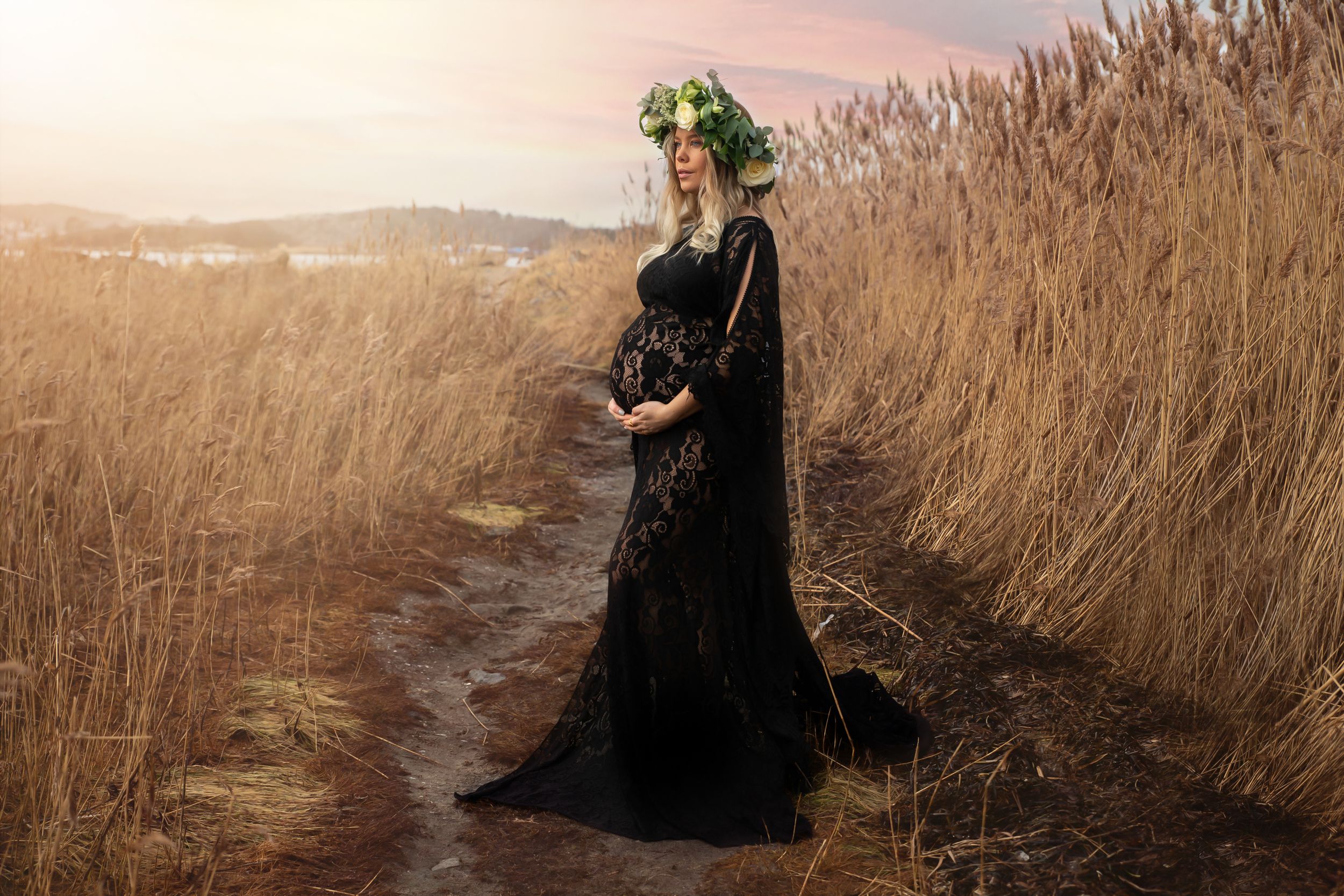 Maternity photoshoot featuring woman in flowing black dress and green floral crown among dried reeds.