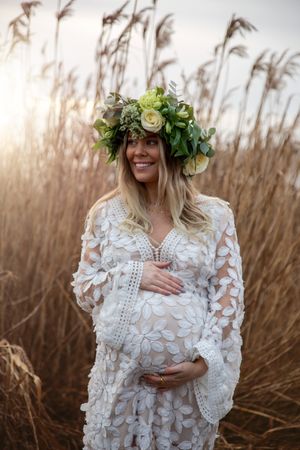Elegant maternity photo with woman in white lace dress and floral crown against golden reeds.