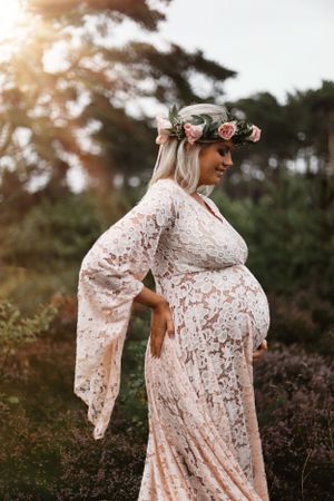 Side profile maternity photograph featuring delicate lace dress and floral crown in natural outdoor setting.